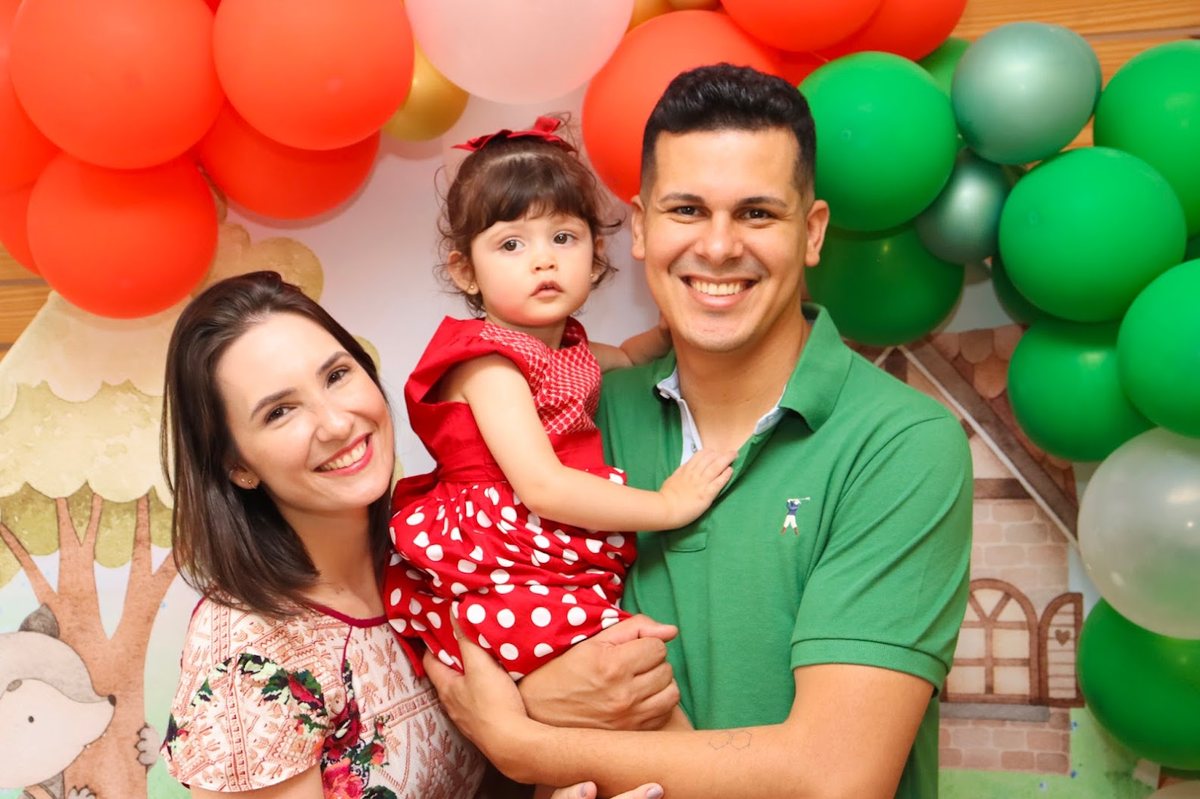 Pai e mãe com a filha no colo, de vestido vermelho de bolinhas, na frente de balões vermelhos e verdes