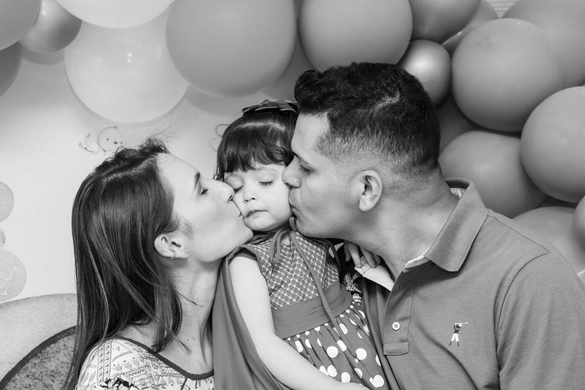 Pai e mãe beijando o rosto da filha ao mesmo tempo, com balões ao fundo, em preto e branco