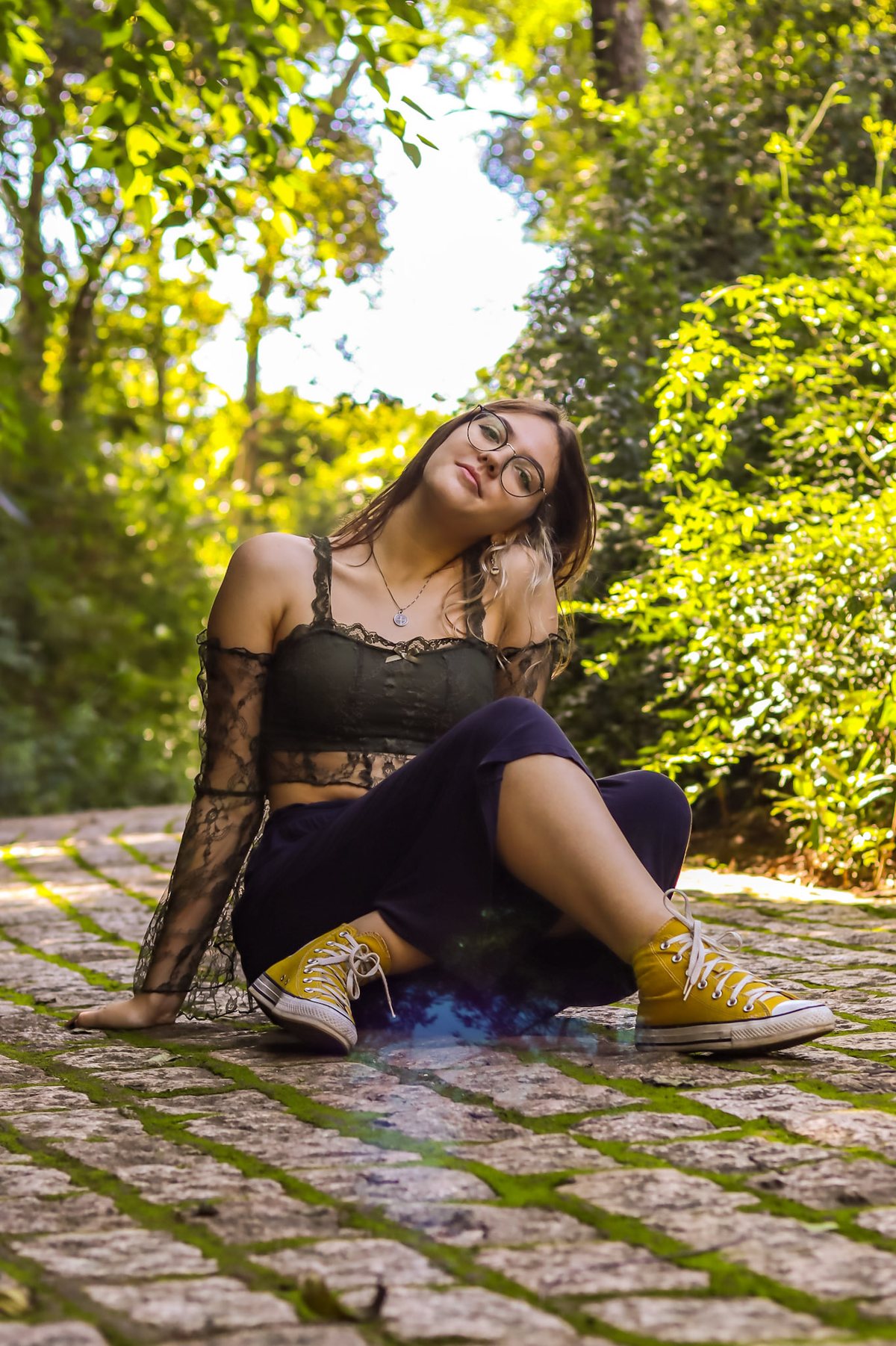 Moça sentada num caminho de pedra no meio das árvores, com luz de sol entre as folhas