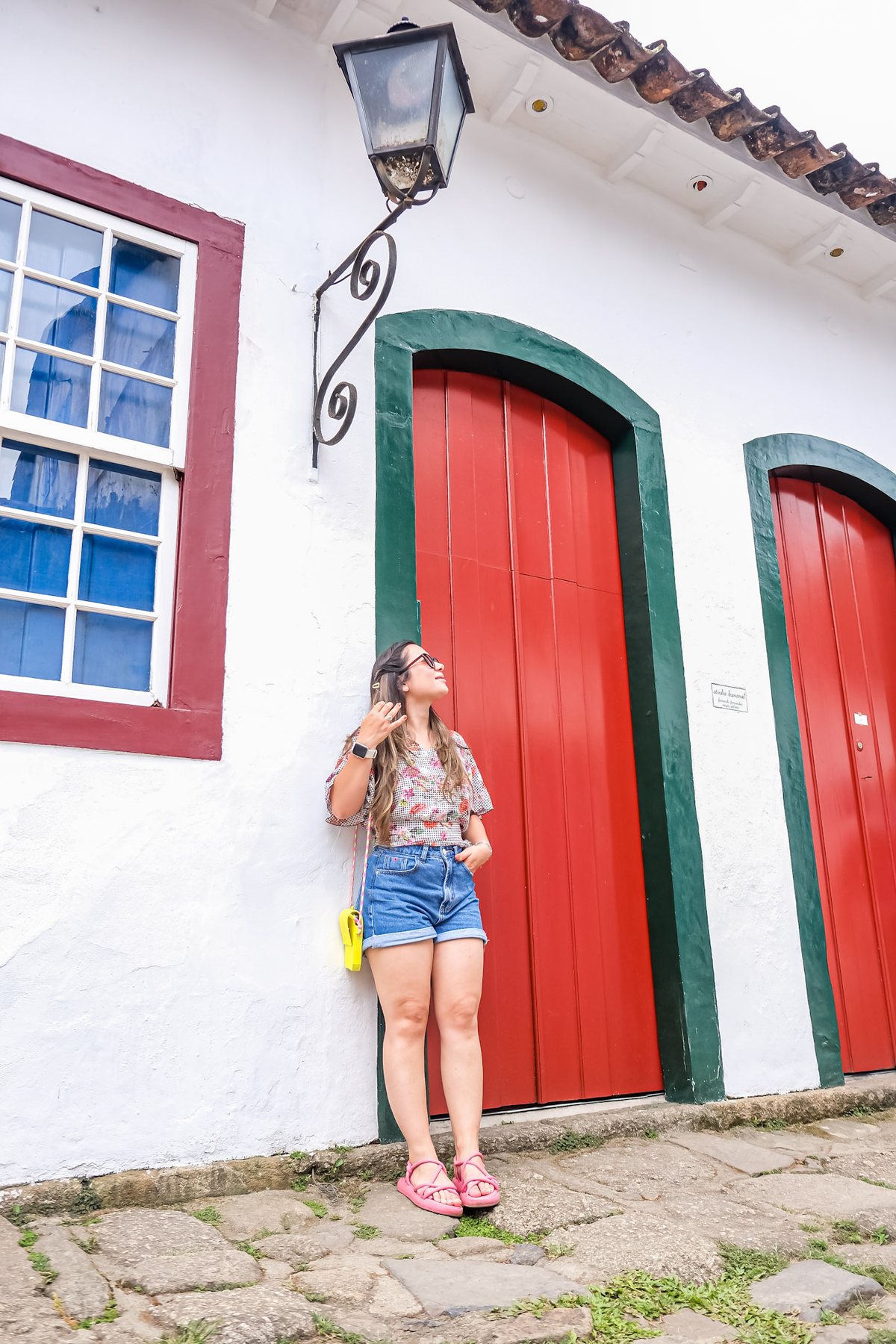 Moça em frente a um casarão branco de portas vermelhas, numa rua de pedra