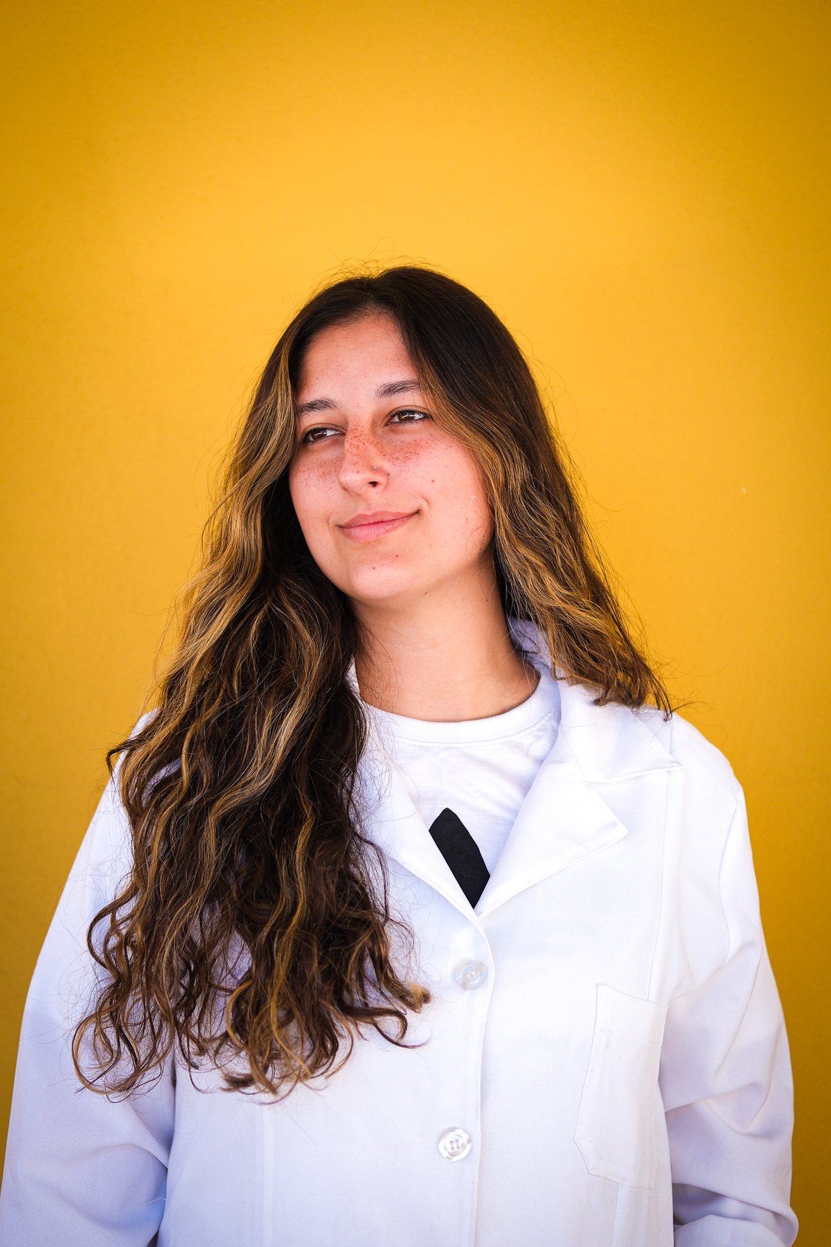 Moça de cabelo longo e jaleco branco, sorrindo, na frente de um fundo amarelo