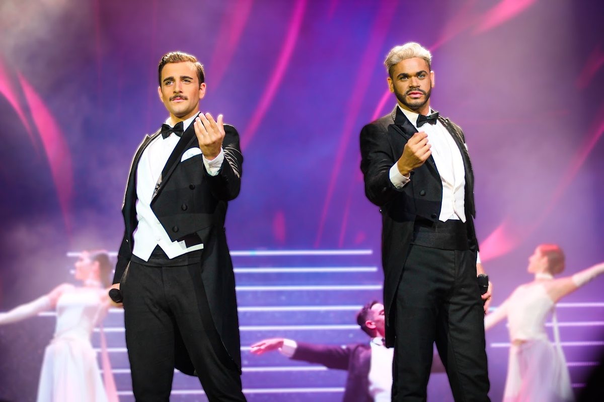 Dois cantores de fraque no palco, com bailarinas ao fundo e luz roxa